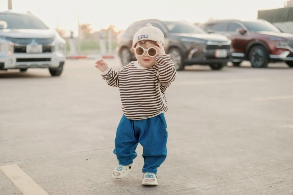 Cute toddler in stylish outfit and sunglasses walking in a parking lot.