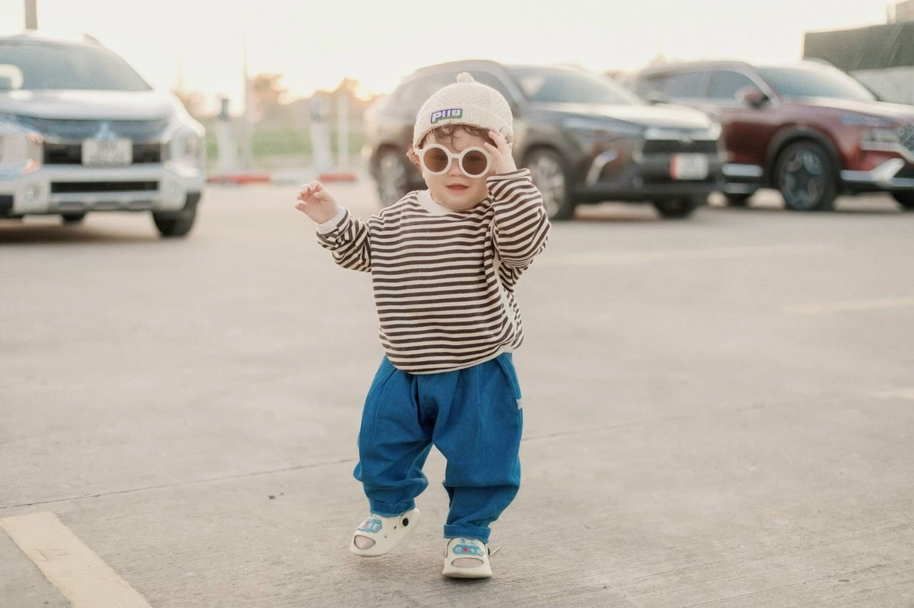Cute toddler in stylish outfit and sunglasses walking in a parking lot.