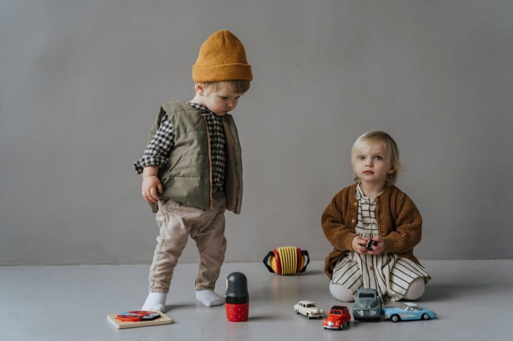 Two fashionable children play with toy cars in an indoor setting, showcasing autumn fashion.